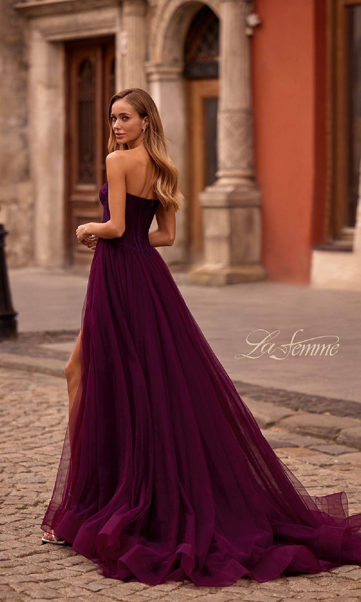 A woman wearing the La Femme 32579 Strapless Long Prom Ball Gown with a floral corset-style bodice and purple floor-length train stands on a cobblestone street, glancing over her shoulder.